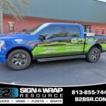 Blue and green wrapped pickup truck advertising B2B Sign & Wrap Resource with mobile sign service and contact info on the side panel.